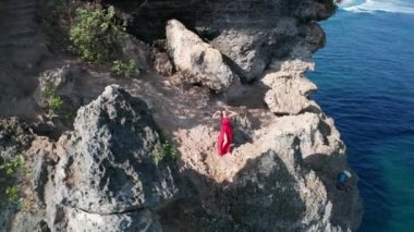 Güneşli yaz gününde deniz kıyısındaki kayalıkta poz veren kırmızı elbiseli güzel kadının havadan drone görünümü