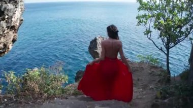 Güneşli yaz gününde deniz kıyısında ki kayalıkta yürüyen kırmızı elbiseli güzel kadının havadan drone geri görünümü