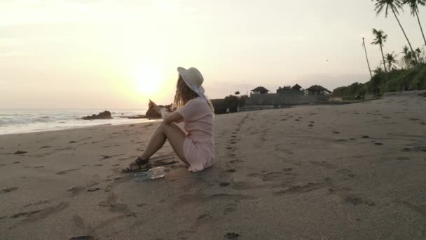 美しい夏の夕日の間に砂浜でリラックスドレスと帽子のかなり幸せな女の子 — ストック動画