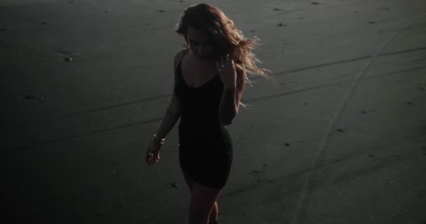 Smyslná brunetka dívka v černých šatech a boty pózující na černé písečné pláži - video ve zpomaleném filmu