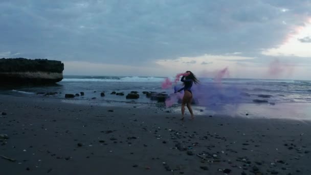 性感的黑发女孩在紫色的体衣冒充彩色烟雾在黑色沙滩在日落 — 图库视频影像