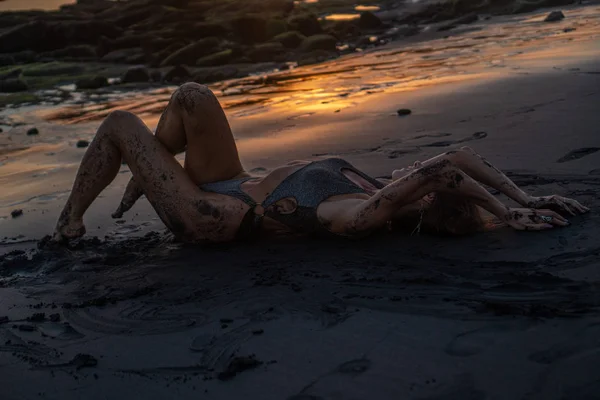 驚くべき黄金の夕日の間に黒い砂浜でポーズキラキラ水着で美しい官能的な女性 — ストック写真