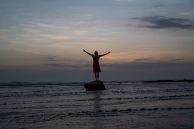 Gün batımında deniz suyundaki küçük kayanın üzerinde poz veren kırmızı elbiseli kadının silueti 
