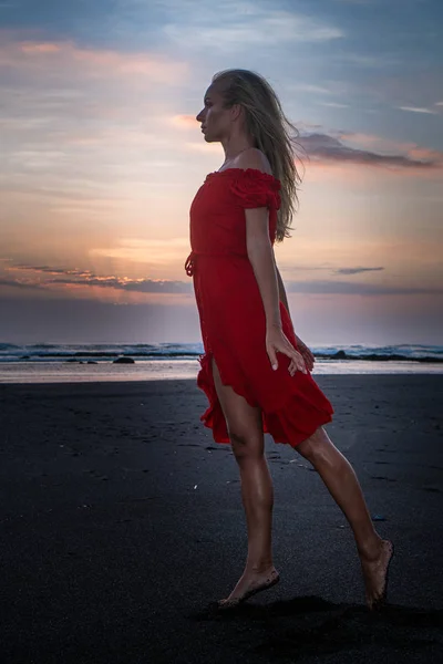 日没時に黒い砂浜でポーズをとる赤いドレスを着た美しい官能的な女性 — ストック写真