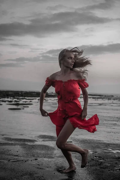 日没時に黒い砂浜を走る赤いドレスを着た美しい官能的な女性 — ストック写真
