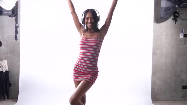 快乐的年轻亚洲女孩在礼服与耳机跳舞在白色的墙壁背景 — 图库视频影像