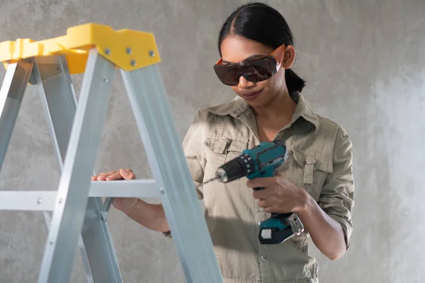 有吸引力的年轻女孩建设者的肖像与钻在整体和保护眼镜站在梯子上混凝土墙背景 — 图库照片