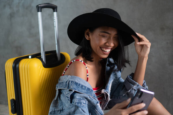Portrait of smiling young Asian woman with luggage doing selfie photo with her mobile phone isolated over concrete wall. People lifestyle, travel concept