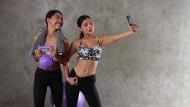 Spor salonunda selfie çekerken spor topu ve dambılla poz veren iki güzel Asyalı kadın. Mutlu bayan arkadaşlar boş zamanlarını spor merkezinde geçiriyorlar.