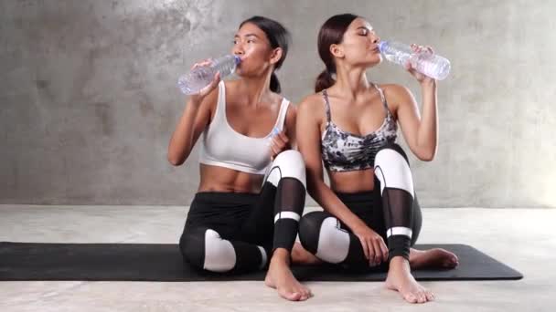 2人の美しい若いアジアの女性が休憩を取り ジムのヨガマットで水を飲んでいます 幸せな女性の友人はスポーツセンターで彼らの自由な時間を過ごす — ストック動画