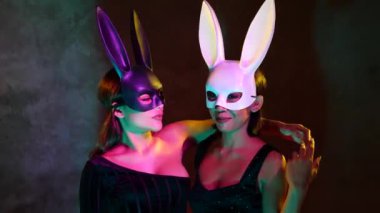 Siyah beyaz tavşan maskeli sevimli Asyalı iki genç kadının portresi pembe ve sarı neon ışıklarda kamera önünde şakacı bir şekilde poz veriyor.