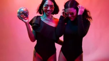 Siyah takım elbiseli, güneş gözlüklü, yakışıklı Asyalı iki genç bayan renkli parlak ışıklarda kamera için dans ediyorlar.