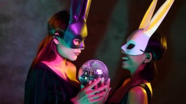 Siyah-beyaz tavşan maskeli, sevimli Asyalı iki genç kadının portresi neon ışıklarda küçük disko topuyla kameraya neşe içinde poz veriyor.
