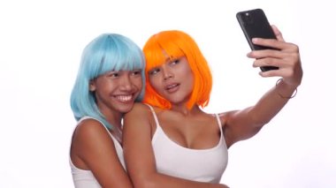 Parlak mavi ve turuncu peruklu iki güzel Asyalı kadının portresi. Beyaz arka planda akıllı telefonlu selfie çekiyorlar.