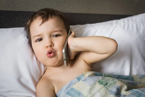 Unga liten pojke prata mobiltelefon ligger på sängen i morgon — Stockfoto