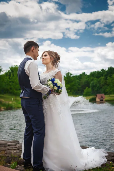 Счастливая невеста и жених улыбаются на природе возле озера с фонтаном — стоковое фото