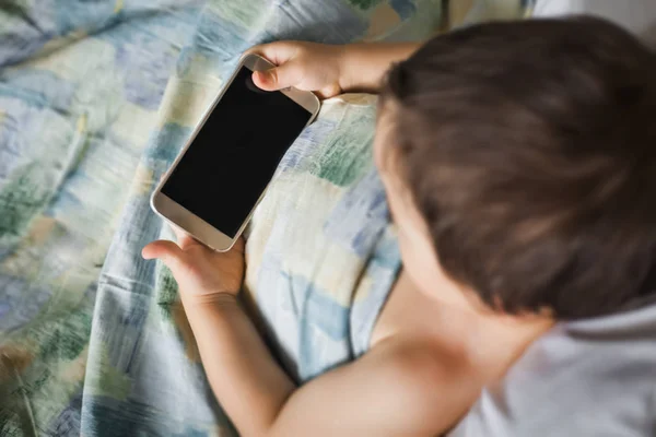 Modern liten pojke håller telefonen ligger på sängen i morgon — Stockfoto