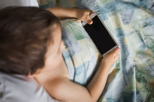 Маленький мальчик держит трогательный телефон лежащий на кровати утром — стоковое фото