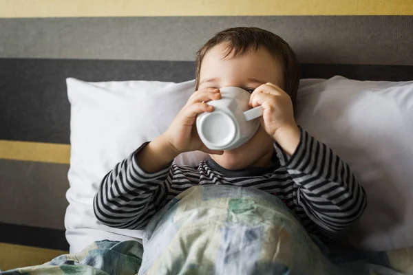 Unwell small boy drinking cup of medicament lying on bed — Stock Photo, Image