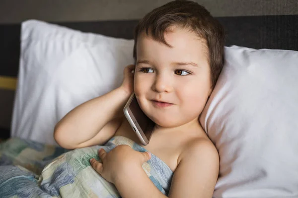Щасливий маленький хлопчик говорить мобільний телефон лежить на ліжку вранці — стокове фото