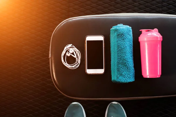 Telefone celular, gotas de ouvido, toalha e uma coqueteleira em um banco em um ginásio — Fotografia de Stock