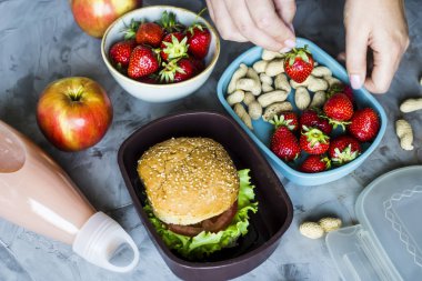 Çocuk okula için öğle yemeği yemek. Gri mutfak masasının üstünde. Sandviç, çilek ve beslenme çantaları fıstık. Üstten Görünüm. Kadın çilek bir kap içinde koyar.
