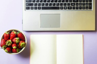 Bahar işyeri dizüstü bilgisayar, dizüstü bilgisayar ve bir kase çilek mor bir arka plan üzerinde. Üstten görünüm, düz yatıyordu