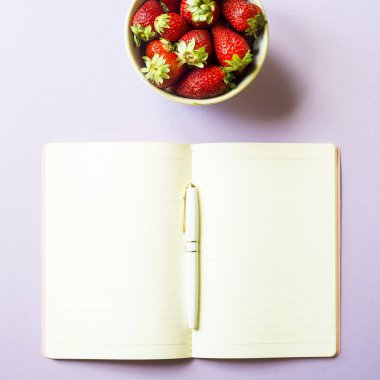 Olgun kırmızı çilek bir kâse mor bir arka plan üzerine açık bir not defteri yanında duruyor. Bahar kavramı. Minimalizm. Üstten görünüm, düz yatıyordu