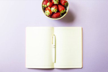 Olgun kırmızı çilek bir kâse mor bir arka plan üzerine açık bir not defteri yanında duruyor. Bahar kavramı. Minimalizm. Üstten görünüm, düz yatıyordu