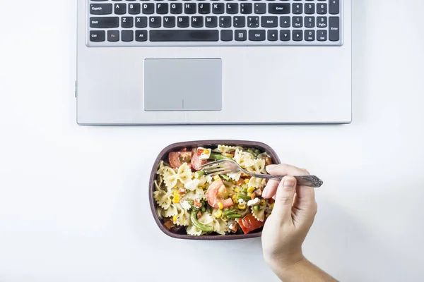 Container with salad with pasta in the workplace near the computer. Lunch in the office during a break between work. Woman take a food. Top view, flat lay