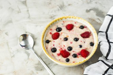Bir tabak yulaf ezmesi ile ahududu ve yaban mersini beyaz kareli peçete yanındaki gri bir tablo. Sağlıklı yaz kahvaltı