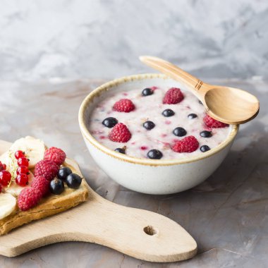 Bir tabak yulaf ezmesi fıstık ezmesi ve banana, sporodina, ahududu sandviçle kurula yanında çilek ile. Sağlıklı yaz kahvaltı gri bir tablo