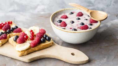 Bir tabak yulaf ezmesi fıstık ezmesi ve banana, sporodina, ahududu sandviçle kurula yanında çilek ile. Sağlıklı yaz kahvaltı gri bir tablo