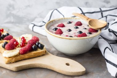 Bir tabak yulaf ezmesi fıstık ezmesi ve banana, sporodina, ahududu sandviçle kurula yanında çilek ile. Sağlıklı yaz kahvaltı gri bir tablo