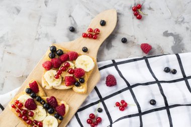 Fıstık ezmesi, muz ve ahududu ve gri bir tablo üzerinde bir ahşap tahta üzerinde kuş üzümü ile Sendvci. Vegan sağlıklı kahvaltı