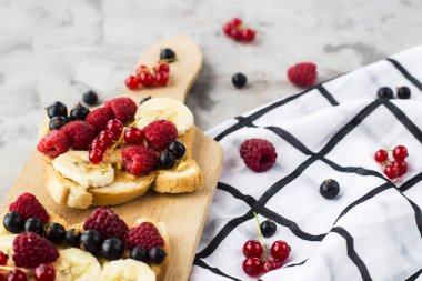 Fıstık ezmesi, muz ve ahududu ve gri bir tablo üzerinde bir ahşap tahta üzerinde kuş üzümü ile Sendvci. Vegan sağlıklı kahvaltı