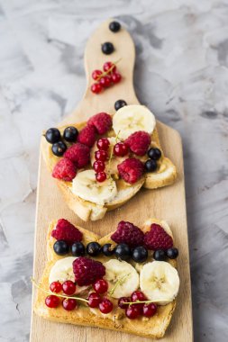Fıstık ezmesi, muz ve ahududu ve gri bir tablo üzerinde bir ahşap tahta üzerinde kuş üzümü ile Sendvci. Vegan sağlıklı kahvaltı