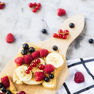 Fıstık ezmesi, muz ve ahududu ve gri bir tablo üzerinde bir ahşap tahta üzerinde kuş üzümü ile Sendvci. Vegan sağlıklı kahvaltı