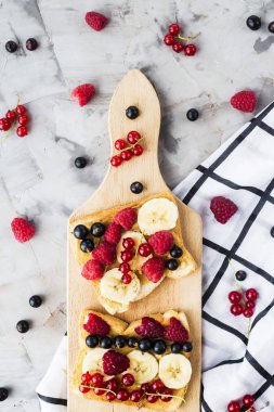 Fıstık ezmesi, muz ve ahududu ve gri bir tablo üzerinde bir ahşap tahta üzerinde kuş üzümü ile Sendvci. Vegan sağlıklı kahvaltı. Üstten görünüm, düz yatıyordu