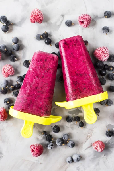 Berry ice cream on a stick of raspberries and currants, homemade with the help of special molds. On a gray table among frozen berries. Top view, flat lay