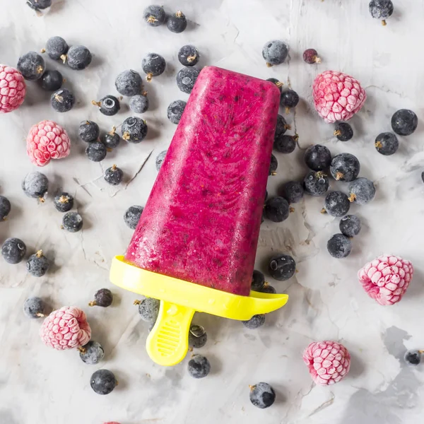 Berry ice cream on a stick of raspberries and currants, homemade with the help of special molds. On a gray table among frozen berries. Top view, flat lay