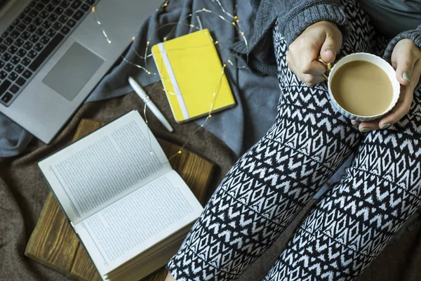A woman is sitting on a rug next to a laptop and a notebook for notes with a cup of coffee. Working in a cozy home environment in winter or autumn
