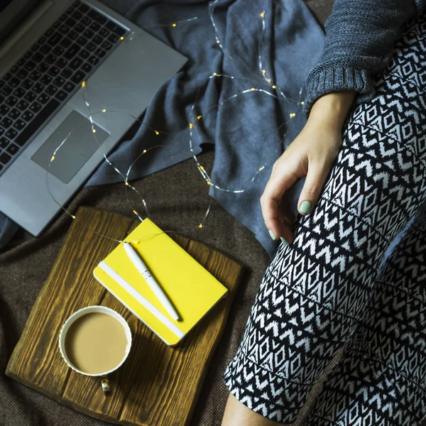 A woman is sitting on a rug next to a laptop and a notebook for notes with a cup of coffee. Working in a cozy home environment in winter or autumn