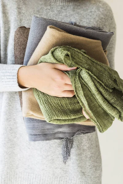 A stack of plaids and scarves in the hands of a woman in a gray sweater. Preparation in cold autumn and winter. Seasonal Wardrobe Concept