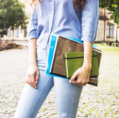 Uzun kıvırcık saçlı bir kız öğrencinin eğitim materyalleri üniversite avlusunda klasörlerde tutuyor. Yüksek öğretim kavramı