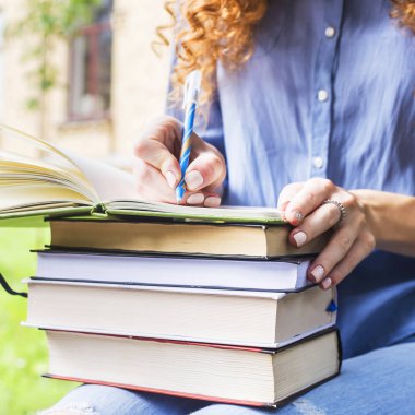 Uzun kıvırcık saçlı kız öğrenci bir defterde bir yığın kitap yazıyor. Üniversitesi'nde ders notları. Çalışma kavramı