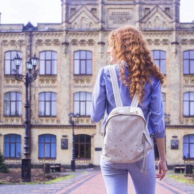 Kızıl saçlı, kıvırcık saçlı öğrenci sırtında bir sırt çantası ile Üniversite binası yanında duruyor. Çalışma için şık moda aksesuar