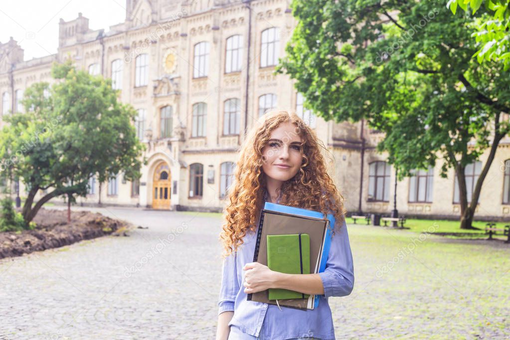 A curly red-haired girl is standing near the university building with educational materials, folders and a notebook for lectures in her hands. Study concept. Back to school