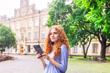 Kıvırcık kırmızı saçlı güzel bir genç kız bir e-kitap tablet onun elinde tutan hangi are için üniversite avluda duran kulaklık bağlı. Çalışma kavramının teknoloji kullanımı