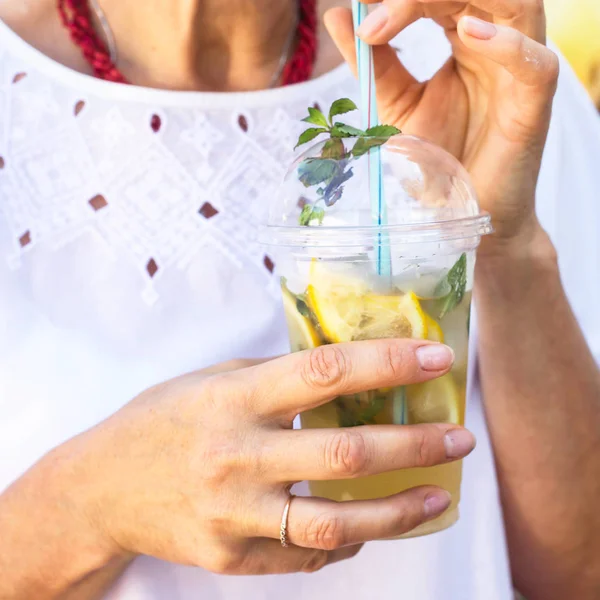 A woman in a white blouse holds a plastic glass with a mojito mug and a mint. Summer drink to cool outside